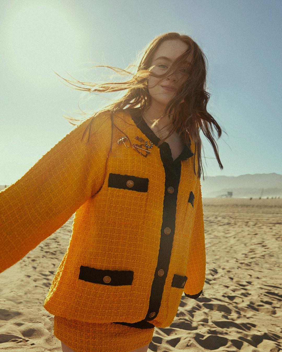 A woman on a sunny beach in a yellow tweed jacket with black trim and short skirt, posing in a carefree manner with the wind in her hair.