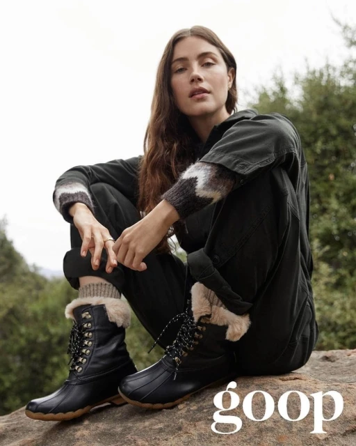 A woman sits on a rock wearing black duck boots with faux fur trim, dark clothing, and neutral-toned socks, in a bright outdoor setting.