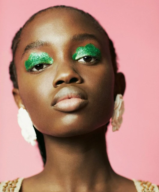 Close-up of a young Black woman with bright green glitter eyeshadow and statement white earrings against a light pink background.