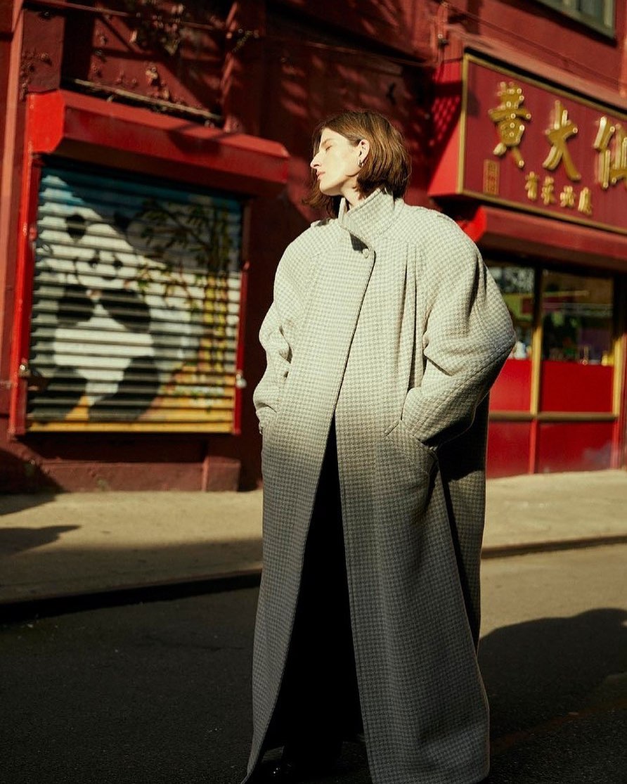 Adult female posing in a light gray houndstooth oversized coat on a city street, in front of a building with Asian signage.
