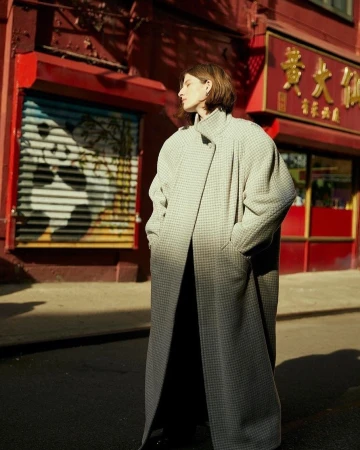 Adult female posing in a light gray houndstooth oversized coat on a city street, in front of a building with Asian signage.