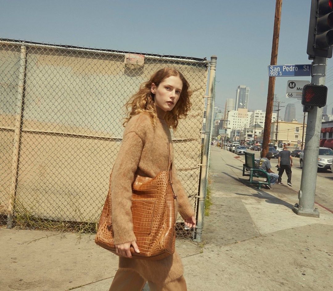 Woman in a brown outfit walks on a city sidewalk carrying a brown reptile-skin tote bag, with buildings in the background on a sunny day.