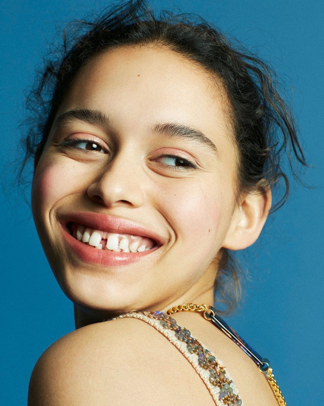 Portrait of a young woman with gap teeth smiling against a blue background, showcasing her shoulder, collarbone, and an ornate strap.