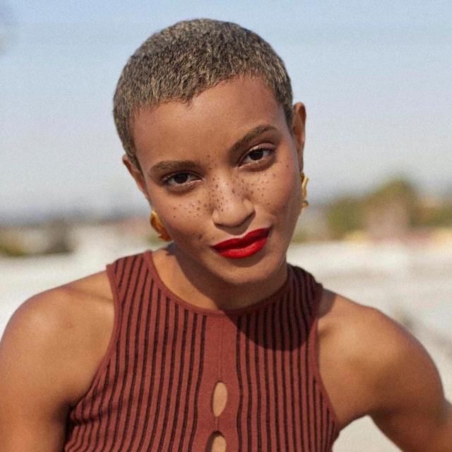 Portrait of a woman with short hair and red lipstick wearing a brown striped top outdoors with a blurred background.