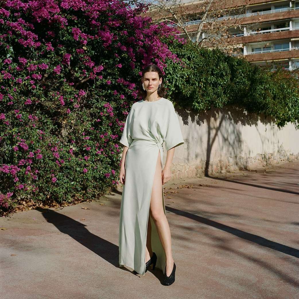 Elegant adult woman in a pale green dress with a thigh-high slit, standing outdoors on a paved surface near a lush bougainvillea bush.
