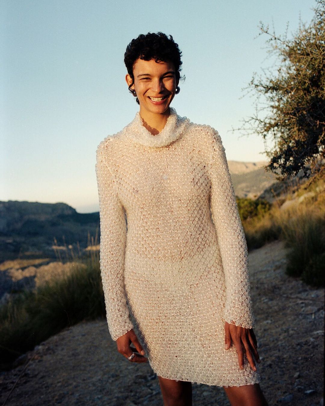 Woman in a textured knit dress stands on a hillside with mountains in the background, smiling at the camera in natural light.