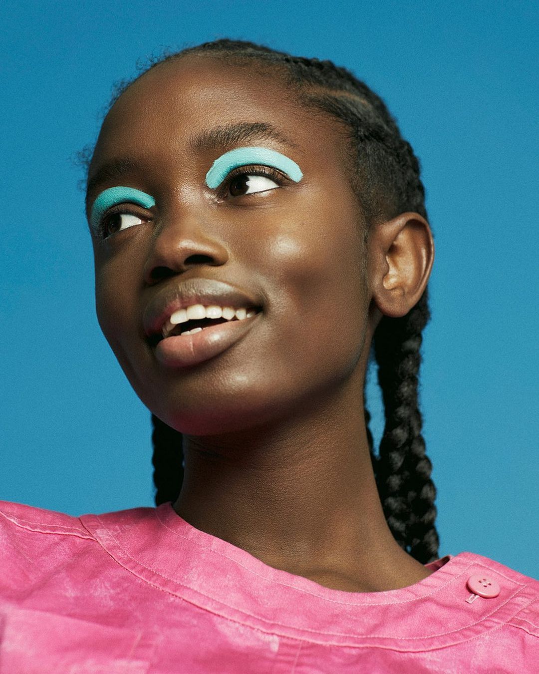 Close-up of a woman with dark skin and braided hair wearing a pink shirt and aqua eyeshadow on a blue background looking away.