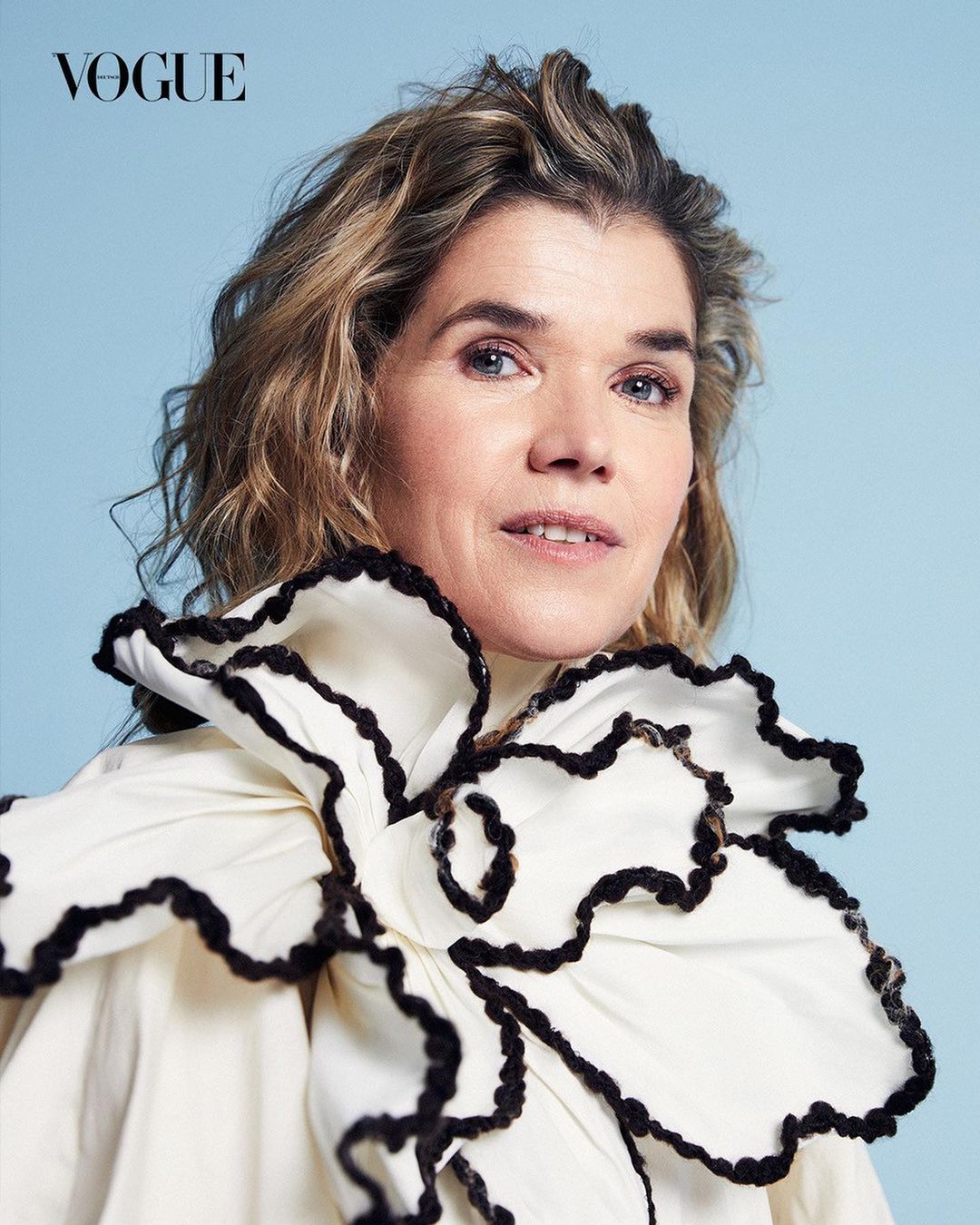 Close-up studio shot of an adult woman with wavy blonde hair wearing a cream-colored top with a large floral ruffled neckline, Vogue logo visible.