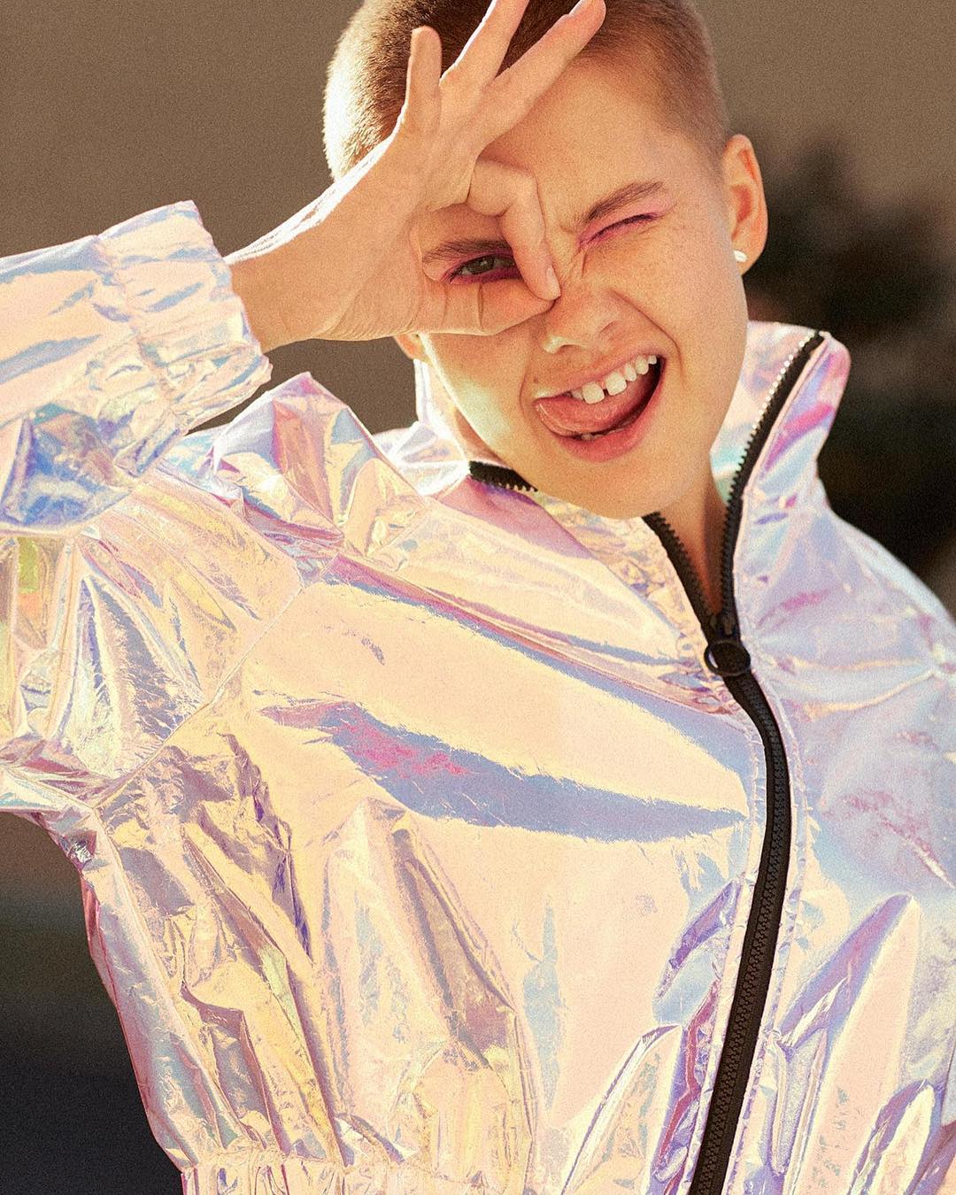 A young, grinning woman with short hair making a circle gesture around her eye and sticking her tongue out, wearing a shiny jacket.