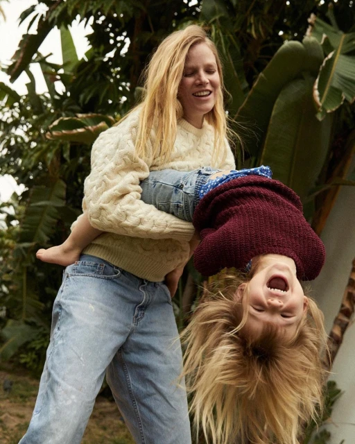 Mother and child play outdoors. Woman holds smiling child upside down. Both are fair-skinned with blond hair and casual clothing.