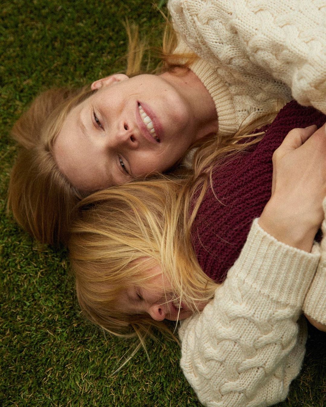 Two smiling women with blonde hair and pale skin lie on a green lawn wearing knit sweaters, viewed in a portrait-style close-up.