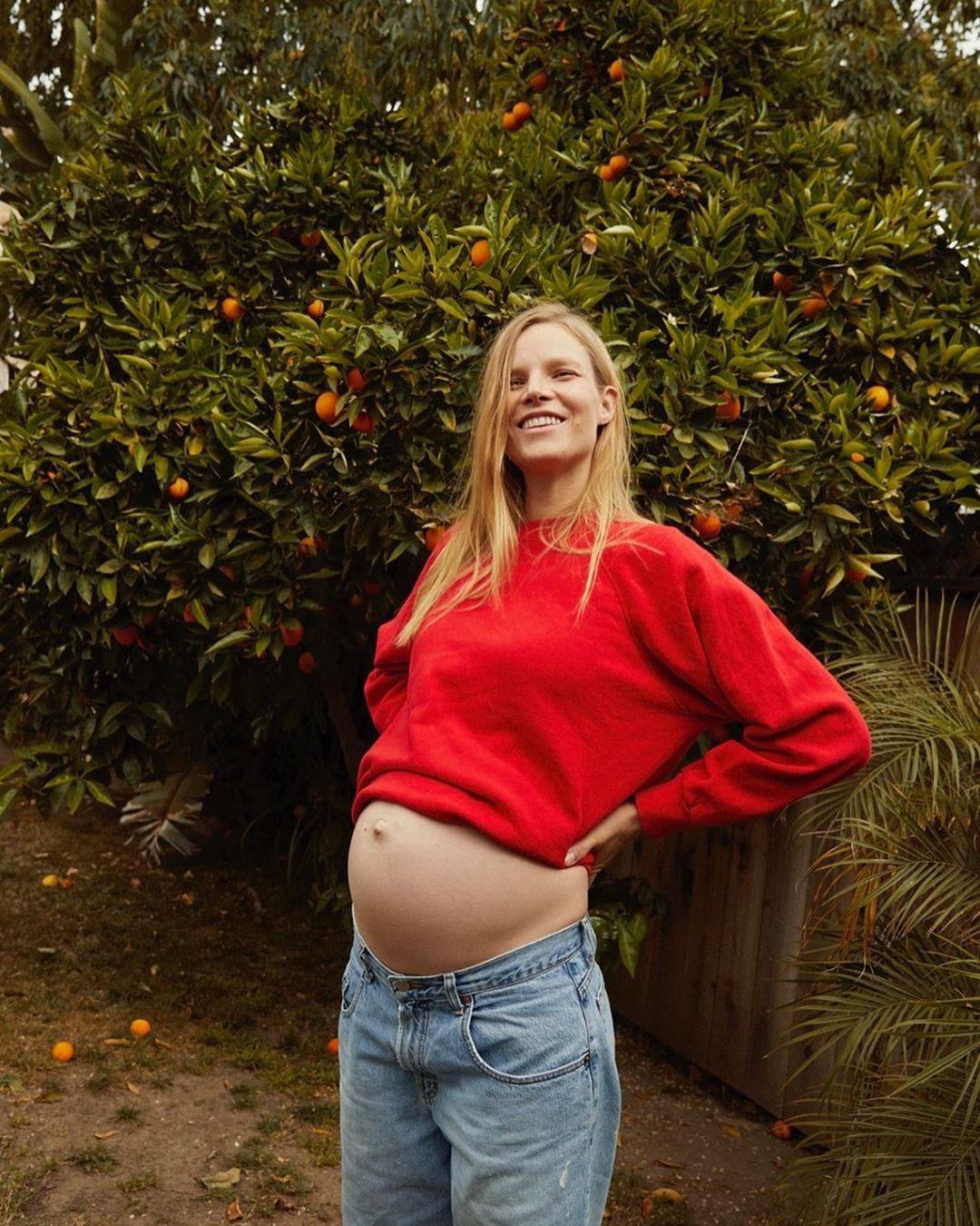 A pregnant adult female stands outside in front of an orange tree, wearing a red shirt and blue jeans, smiling and looking at the camera.