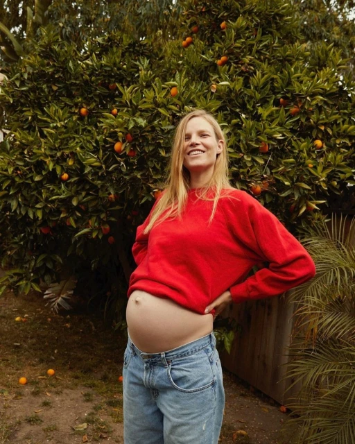 A pregnant adult female stands outside in front of an orange tree, wearing a red shirt and blue jeans, smiling and looking at the camera.