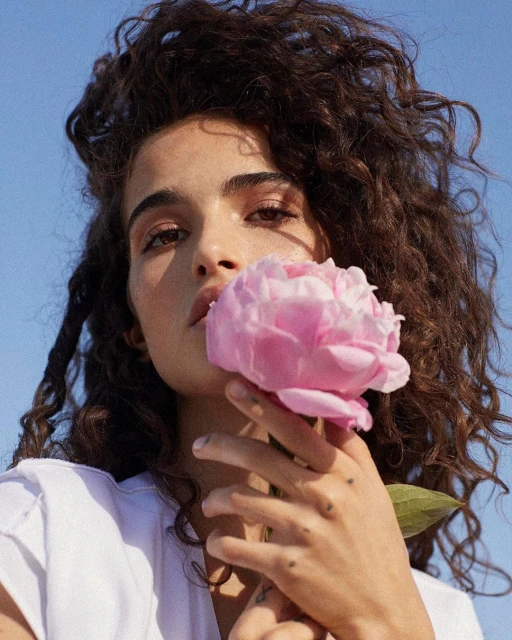 Close-up portrait of a young woman with curly hair holding a pink flower against a blue sky backdrop in natural light.