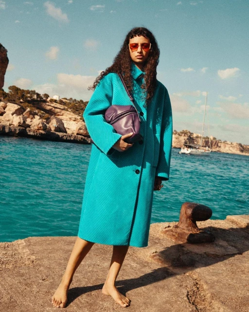 A barefoot woman in an aqua coat, orange sunglasses, and a purple bag poses on a stone surface with a boat and coastline background.