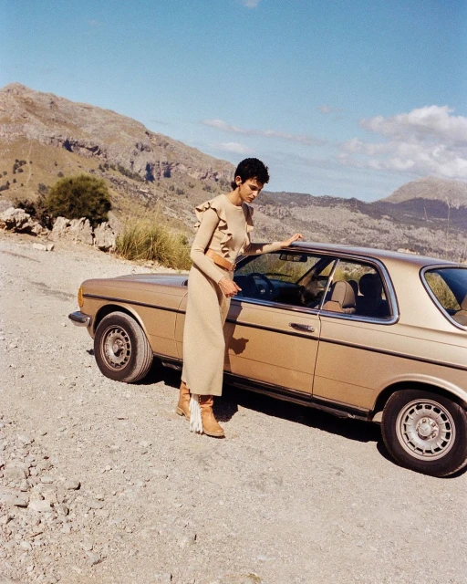 Fashion shot of a woman posing next to a vintage car in a mountainous outdoor setting under a clear blue sky, wearing a dress and boots.
