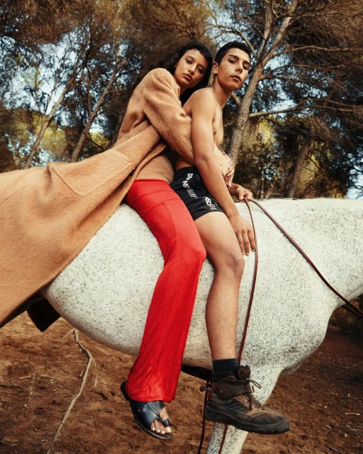 A stylish couple poses with a white horse statue in a sunlit outdoor setting. The woman wears a coat, and the man sports shorts.