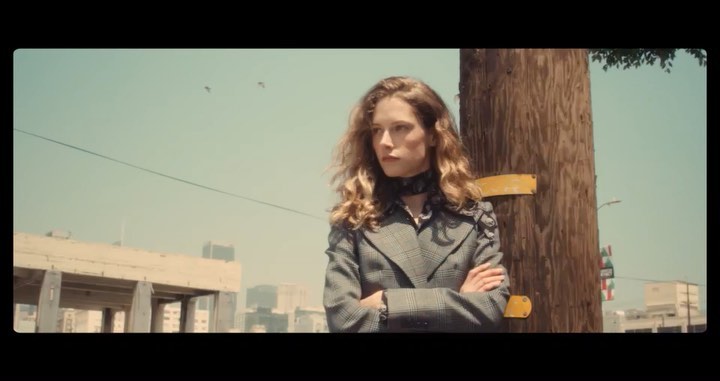 Adult female model in a grey plaid blazer standing in an outdoor urban environment with buildings in the background on a sunny day.