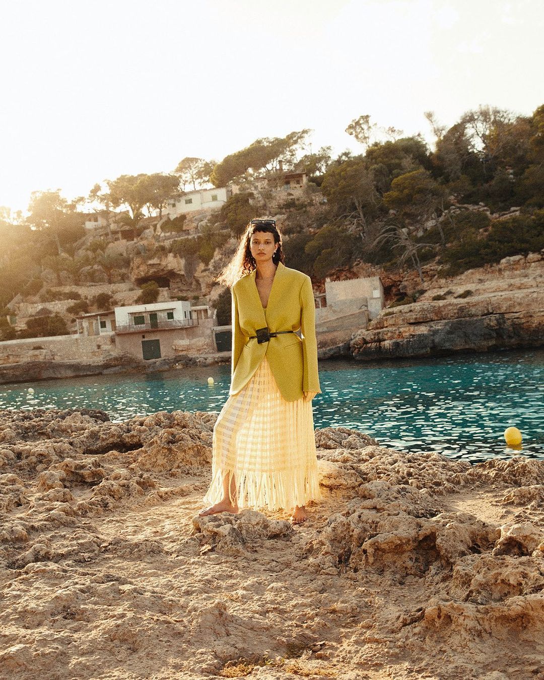 Woman in an oversized yellow blazer and long fringe skirt standing on a rocky outcrop by the sea on a sunny day.