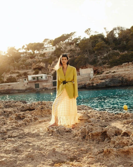Woman in an oversized yellow blazer and long fringe skirt standing on a rocky outcrop by the sea on a sunny day.