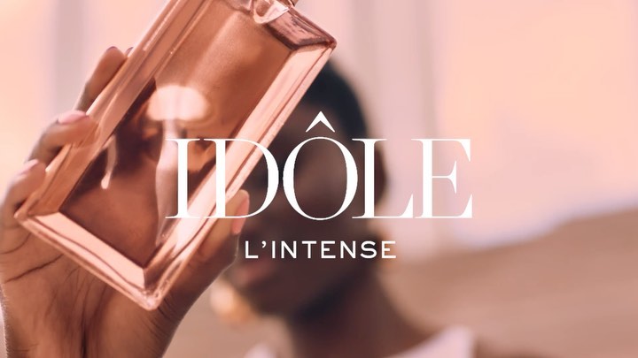 Close-up studio shot of a hand holding a Lancôme Idôle L'Intense perfume bottle against a soft pink backdrop.
