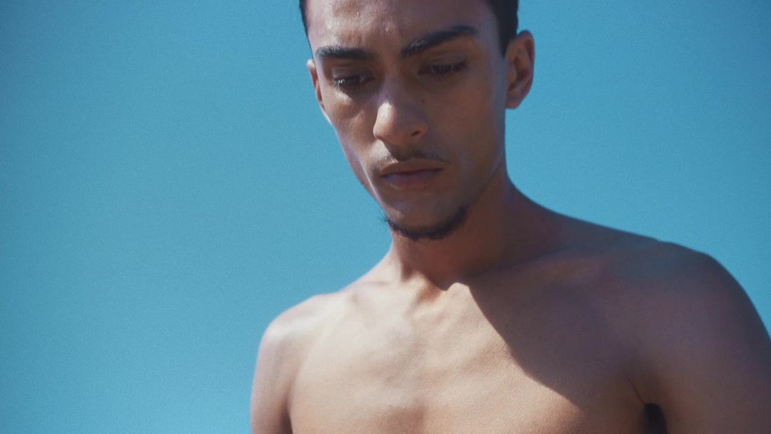 Portrait of a shirtless young man looking down against a bright blue sky background captured by photographer Camilla Armbrust.