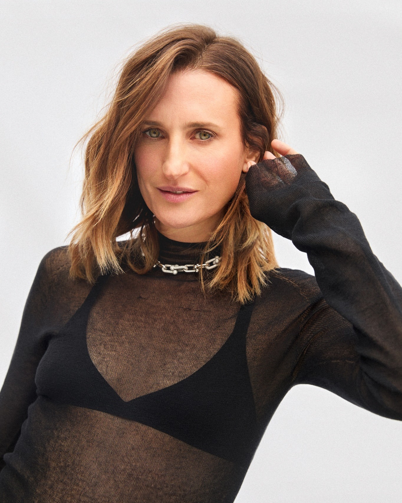 Woman with shoulder-length brown hair wearing a sheer black top and silver chain necklace against a light background.