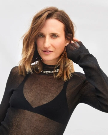 Woman with shoulder-length brown hair wearing a sheer black top and silver chain necklace against a light background.
