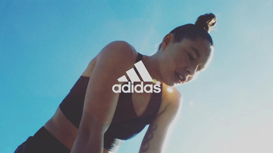A low-angle shot showcases an adult woman with a bun hairstyle, fair skin, and Adidas branding, set against a bright blue sky backdrop.