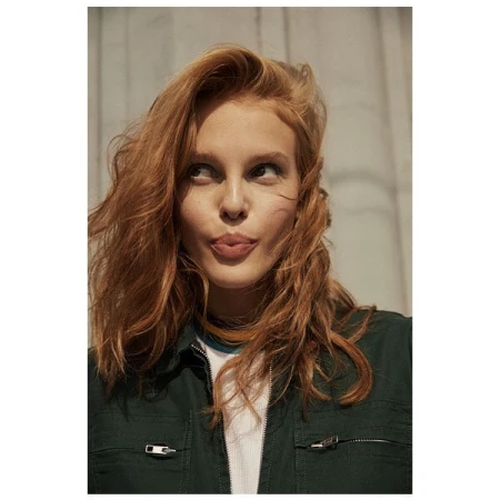 Close-up portrait of a red-haired woman in a green shirt looking to the side, with a neutral expression and light grey background.