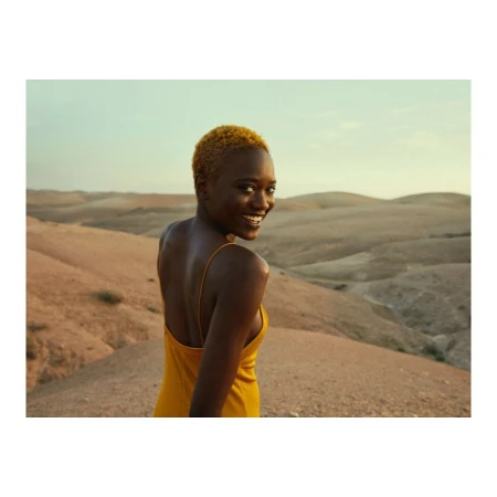 Portrait of a Black woman with short yellow hair wearing a yellow dress, looking back in a desert landscape.