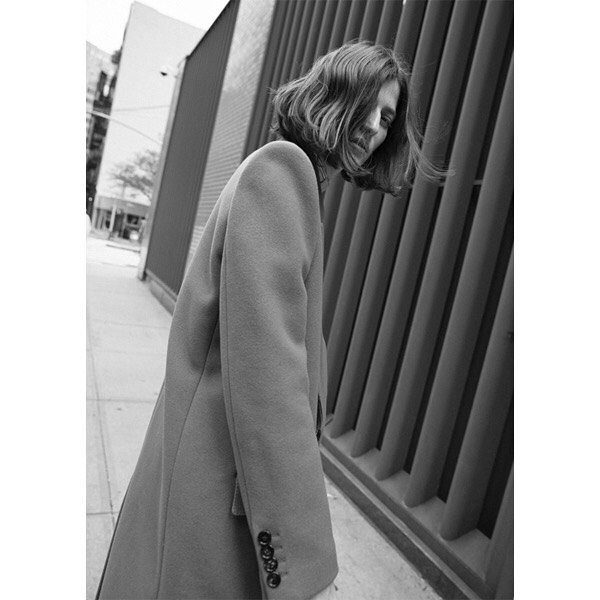 Black and white fashion shot of a woman in a coat walking next to a building on an outdoor city street, leaning into the camera.