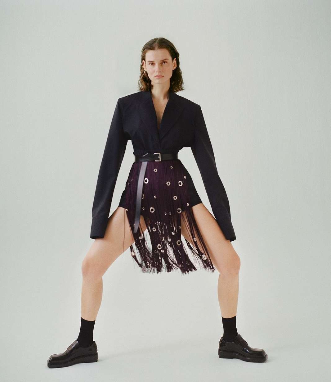Fashionable adult female poses in a dark blazer, distinctive fringe skirt with grommets, black socks, and loafers against a white backdrop.