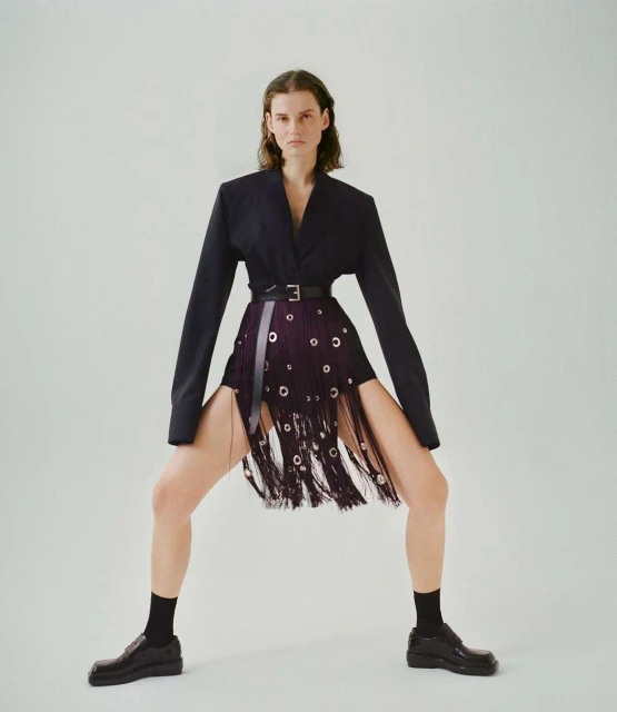 Fashionable adult female poses in a dark blazer, distinctive fringe skirt with grommets, black socks, and loafers against a white backdrop.