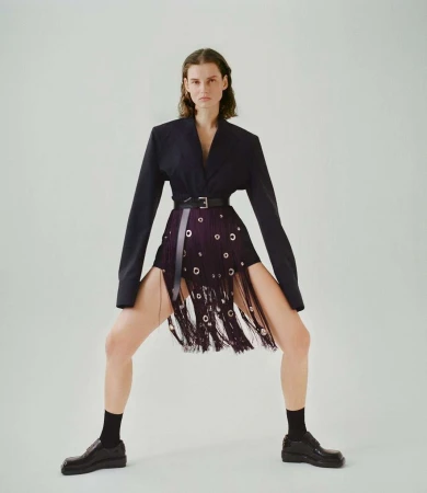 Fashionable adult female poses in a dark blazer, distinctive fringe skirt with grommets, black socks, and loafers against a white backdrop.
