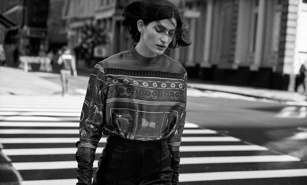 Monochrome outdoor shot of a woman in patterned top and black bottoms crossing a zebra stripe crosswalk with buildings in the background.