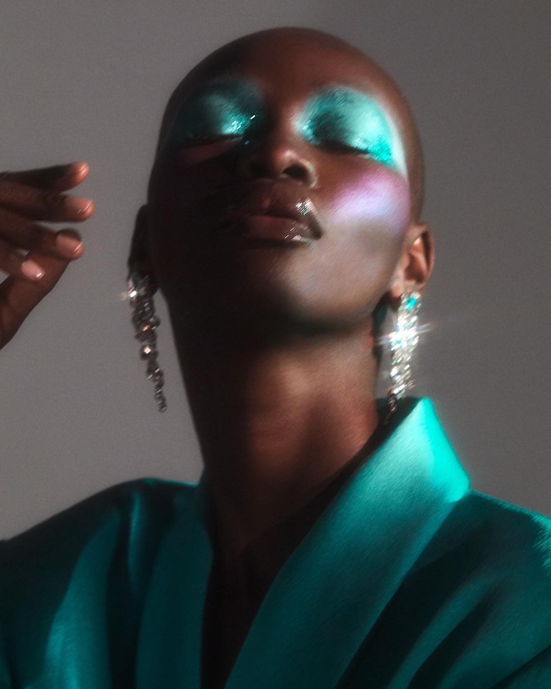 Glamorous bald woman with turquoise makeup and statement earrings poses in a turquoise satin jacket in a studio shot.
