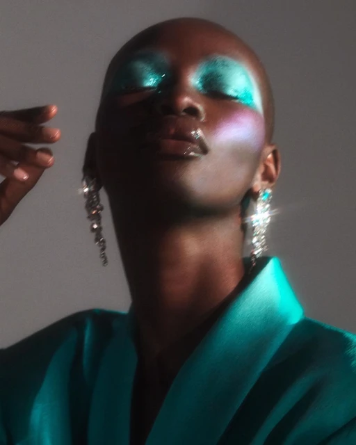 Glamorous bald woman with turquoise makeup and statement earrings poses in a turquoise satin jacket in a studio shot.