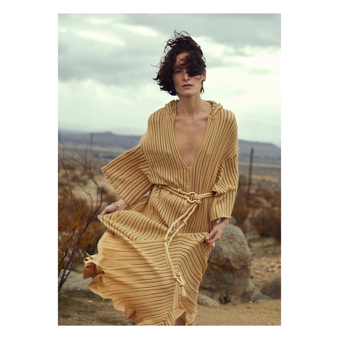 Brunette woman in flowing gold dress with a hood and rope belt poses in a desert landscape under an overcast sky. Fashion shot.
