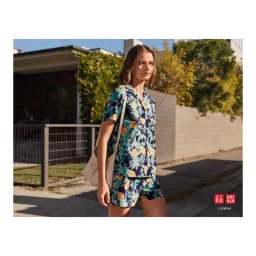 Woman in floral patterned short sleeve shirt and shorts holding shoulder bag outdoors by residential building