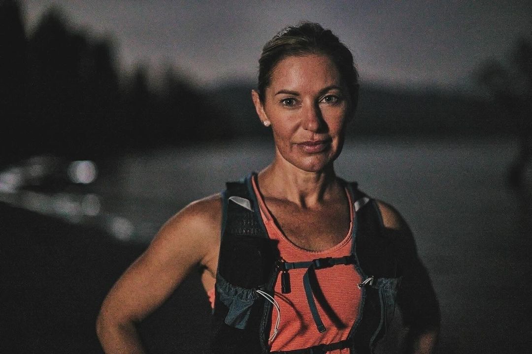 Portrait of an adult woman in fitness wear outside at night with lake and trees in the background, lit by artificial light.