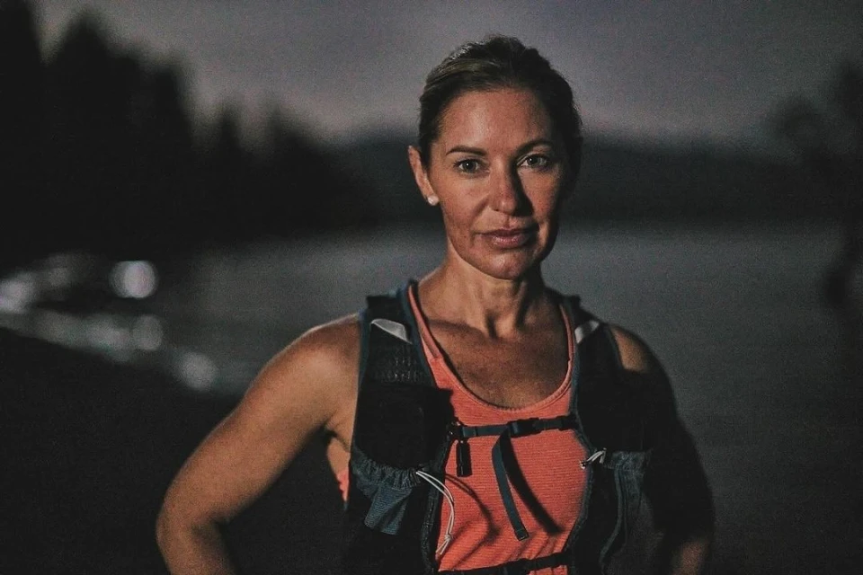 Portrait of an adult woman in fitness wear outside at night with lake and trees in the background, lit by artificial light.