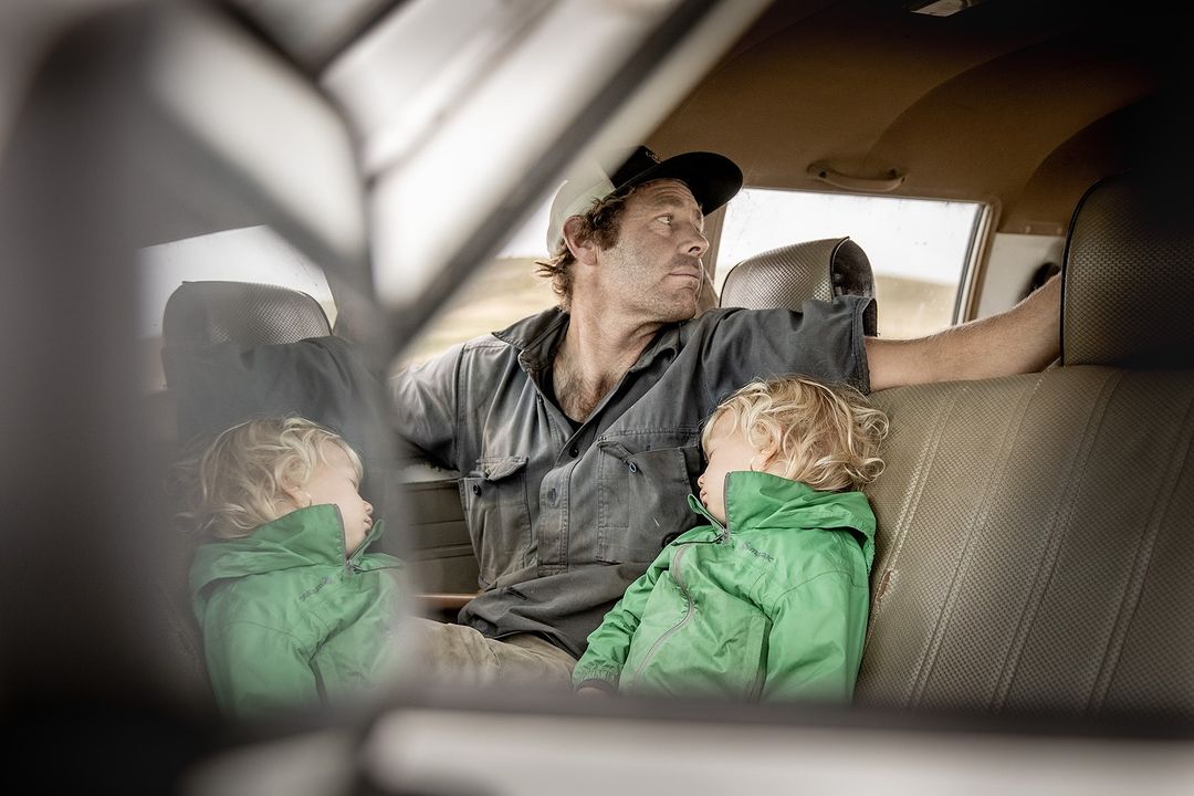 Man and two children inside car, with the children resting, the man looking out the window, and the vehicle's interior visible.