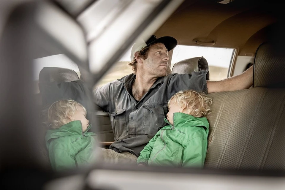 Man and two children inside car, with the children resting, the man looking out the window, and the vehicle's interior visible.