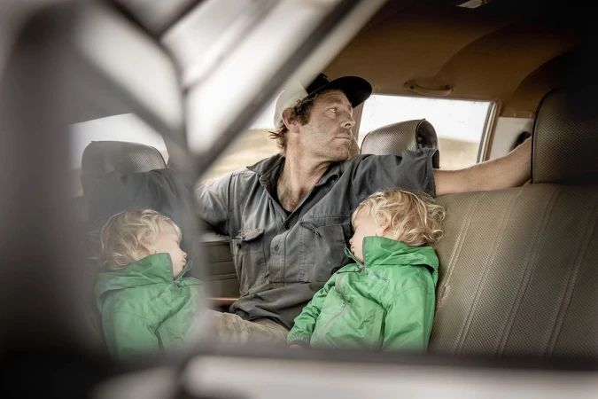Man and two children inside car, with the children resting, the man looking out the window, and the vehicle's interior visible.