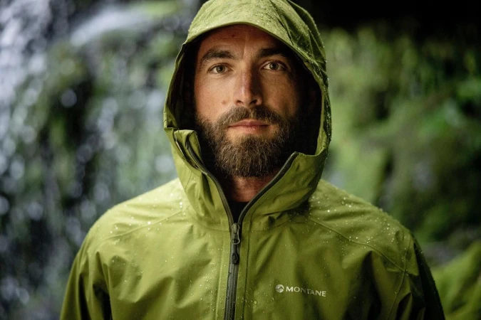 Portrait of a bearded adult male wearing a hooded jacket in a lush outdoor environment, raindrops visible on fabric.