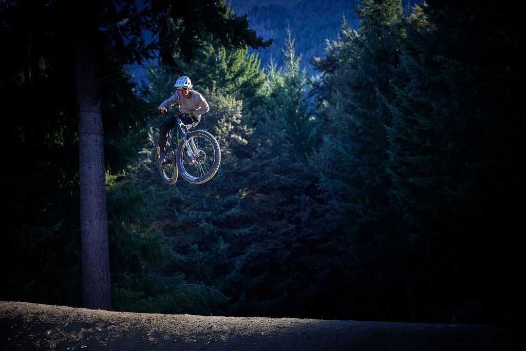 A mountain biker jumps through the air between trees. The scene is dark and shady with a mountain in the background.