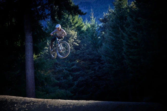 A mountain biker jumps through the air between trees. The scene is dark and shady with a mountain in the background.