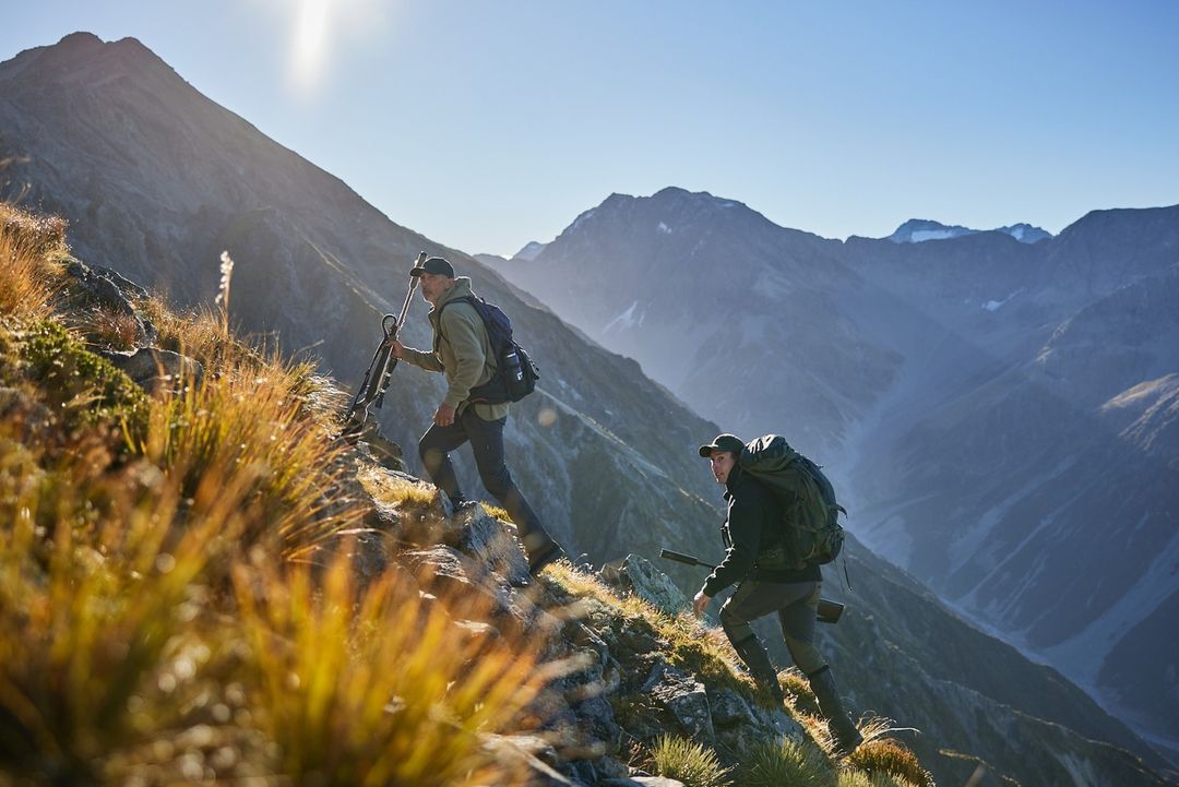 Two men with backpacks and hunting rifles are hiking up a steep mountain ridge in sunny weather, with mountain peaks in the background.