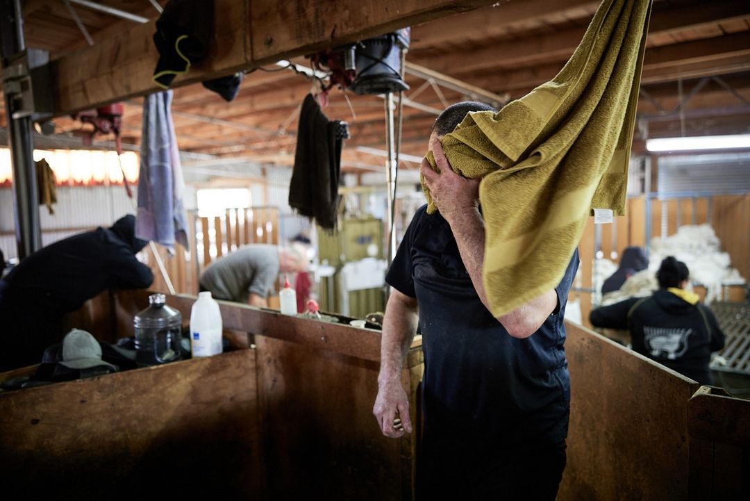 Adult male wiping face with towel inside industrial structure with wooden walls. Other people and supplies are visible in background.